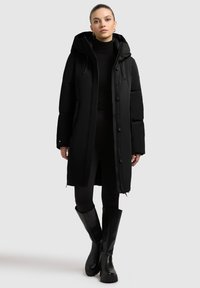 Black puffer coat with a hood, featuring a front button closure, large pockets, and quilted texture; paired with fitted black leggings and knee-high boots.