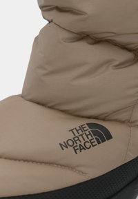 Botte rembourrée beige avec une semelle noire texturée, présentant un logo noir bien visible "The North Face" sur le côté. Matériau lisse et isolant.