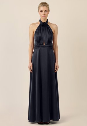 Vrouw draagt een mouwloze, zwarte halterjurk tot op de grond met een geplooide bustier en een roosdetail bij de halslijn, staand tegen een effen achtergrond.