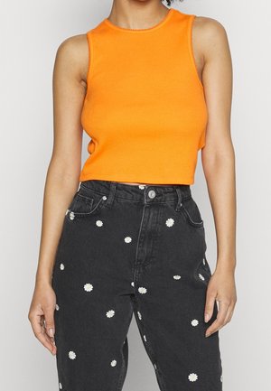 Frau trägt ein ärmelloses, knallorangfarbenes Crop-Top und schwarze, hoch taillierte Jeans mit weißen Gänseblümchenmustern vor einem schlichten Hintergrund.