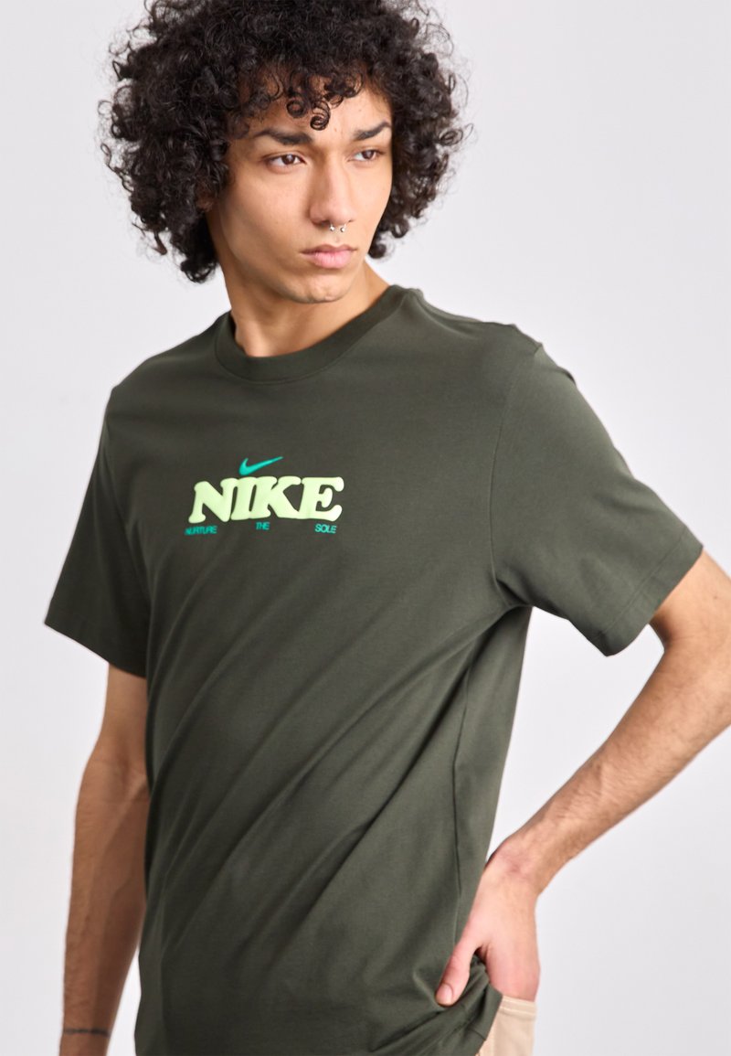 Oliwkowa koszulka Nike wykonana z miękkiej bawełny, z kontrastowym jasnooliwkowym logo i tekstem z przodu; okrągły dekolt, krótkie rękawy.