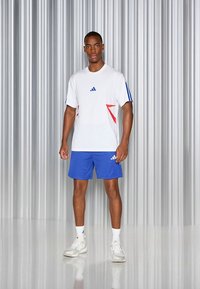 Camiseta desportiva branca com detalhes em vermelho e azul, combinada com calções azuis. Concluída com ténis brancos, sobre um fundo metálico.