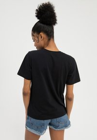 Nainen, jolla on kiharat hiukset nutturalla, pukeutuneena yksiväriseen mustaan lyhythihaiseen t-paitaan ja kuluneisiin farkkushortseihin, katsoo poispäin valkoista taustaa vasten.