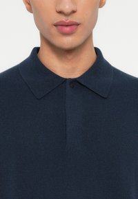 Primo piano di una persona che indossa una polo blu scuro con bottoni, mostrando dettagli del collo, della parte inferiore del viso e del colletto.