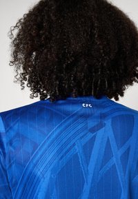 Sininen urheilupaita, jossa on teksturoitu kangas, jossa on vinoviivoja ja pystysuoria kuvioita. "CFC"-logo sijaitsee takana kauluksen kohdalla.