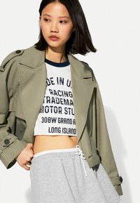 Olijfgroene cropped jas met oversized revers en knoopaccenten, gelaagd over een witte graphic tee en grijze sweatshorts.