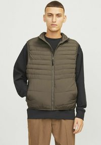 Gepolsterte, olivgrüne Weste mit hohem Kragen, Frontreißverschluss und horizontalem Steppmuster. Getragen über einem schwarzen Langarmshirt.