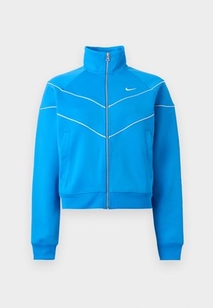 Chaqueta deportiva azul brillante con cremallera, cuello alto, diseño de ribete blanco, bolsillos laterales y pequeño logo blanco de Nike en el pecho.