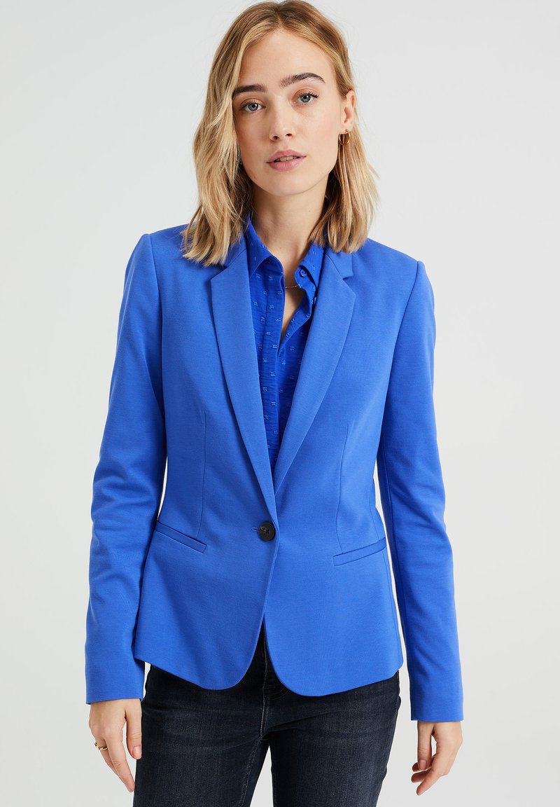 Blauer Blazer aus strukturiertem Stoff, mit Ein-Knopf-Verschluss, Reverskragen und zwei vorderen Taschen, kombiniert mit einem blauen gemusterten Hemd.