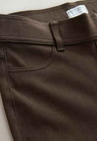 Pantaloni jersey marroni con una texture liscia, caratterizzati da una vita tradizionale, passanti per la cintura e tasche frontali. Sono visibili dei dettagli cuciti.