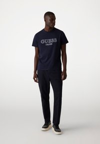 Navyblauw katoenen t-shirt met het 'GUESS' logo in wit, gecombineerd met donkere broeken en zwarte sneakers met een witte zool. Eenvoudig modern ontwerp.