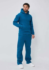 Sweat à capuche bleu sarcelle et pantalon de survêtement assorti en tissu doux. Comprend une poche avant, une capuche à cordon et des poignets côtelés. Porté avec des chaussures blanches.