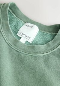 Grøn bomulds sweatshirt med rund halsudskæring og blød tekstur. Etiketten siger "NEXT downtime elevated loungewear." Ren syning synlig.