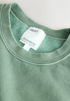 Zielony bawełniany sweter z okrągłym dekoltem i miękką teksturą. Metka mówi "NEXT downtime elevated loungewear". Widoczne czyste szwy.