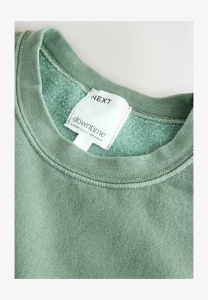 Felpa verde in cotone con scollo rotondo e texture morbida. L'etichetta riporta "NEXT downtime elevated loungewear." Cuciture pulite visibili.