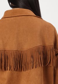 Veste en suède synthétique marron avec un détail frangé le long de l'ourlet arrière et un col, présentant une texture lisse.