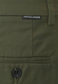 Alyvuogių spalvos kelnės su tekstūruotu audiniu, su juodos spalvos etikete "JACK&JONES" ir tamsiu mygtuku liemenyje.