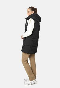Chaleco de plumas negro con capucha, con textura acolchada y detalles en color marrón claro en los hombros. Combinado con pantalones beiges y zapatillas blancas.