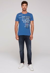Camiseta de algodón azul con cuello redondo, que presenta texto gráfico y números en blanco. Llevada con jeans oscuros y zapatillas blancas.