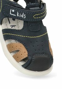 Sandalia azul marino para niño pequeño con costuras amarillas, detalles en marrón, perforaciones y tira de velcro etiquetada "niños" sobre una suela blanca.