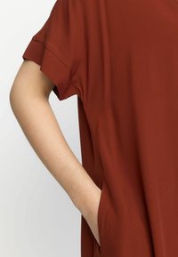 Rostfarbiges Kleidungsstück mit kurzen Ärmeln und lässiger Passform. Die Textur wirkt glatt und verfügt über seitliche Taschen, die in das Design integriert sind.