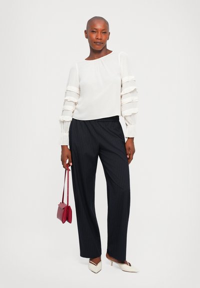 Femme portant une blouse blanche à manches tiered, un pantalon large à rayures noires, des talons pointus blancs, tenant un petit sac à main rouge.