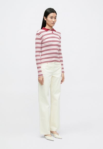 Chemise blanche à manches longues avec des rayures horizontales rouges et un col rouge, associée à un pantalon large crème et des chaussures blanches pointues.