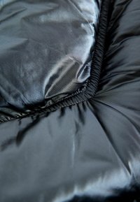 Svart pufferjacka i tyg som visar upp en slät, glänsande yta med kviltade sektioner och elastiska sömmar, vilket betonar isolering och flexibilitet.