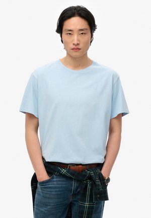 Lichtblauw katoenen t-shirt met korte mouwen, ronde hals en klein logo. Gedragen met donkere jeans en een groen geruite shirt rond de taille geknoopt.