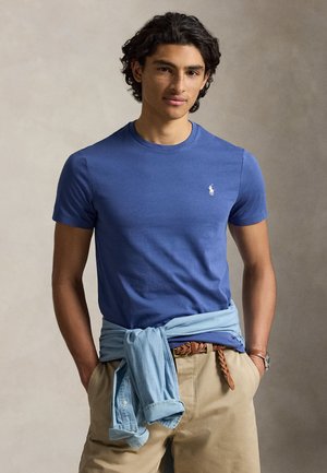 Mlad muškarac s kovrčavom tamnom kosom, odjeven u plavu majicu, bež hlače, svijetloplavu košulju vezanu oko struka i rukama u džepovima.