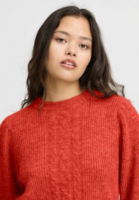Pull en tricot rouge avec un design en torsades au centre. Il a un col rond et un tissu texturé, offrant une apparence chaleureuse.