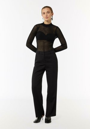 Schwarzes, langärmliges Oberteil aus Mesh mit vertikalen Streifen, kombiniert mit hochtaillierten schwarzen Hosen. Stilettos vervollständigen das Outfit.