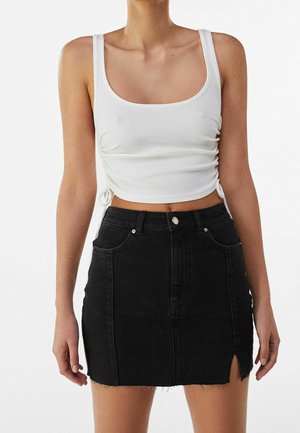 Frau trägt ein weißes, geripptes, kurz geschnittenes Tanktop mit seitlichen Bändern und einen schwarzen, hoch taillierten Jeans-Minirock mit einem Frontschlitz.