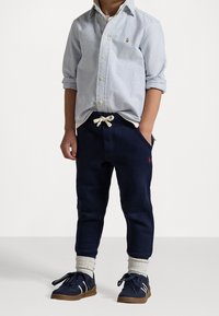 Barn klädd i en blårandig skjorta med uppkavlade ärmar, marinblå joggerbyxor, vita strumpor och marinblå sneakers med bruna sulor.