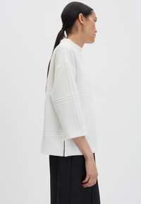 Donna con una lunga coda di cavallo nera che indossa un top bianco con texture a tre quarti e pantaloni neri su uno sfondo neutro.