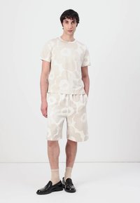 Kortermet beige t-skjorte med blomsterdesign og matchende shorts, laget av myk tekstil. Modellen har på seg lyse sokker og svarte sko. Enkel design.