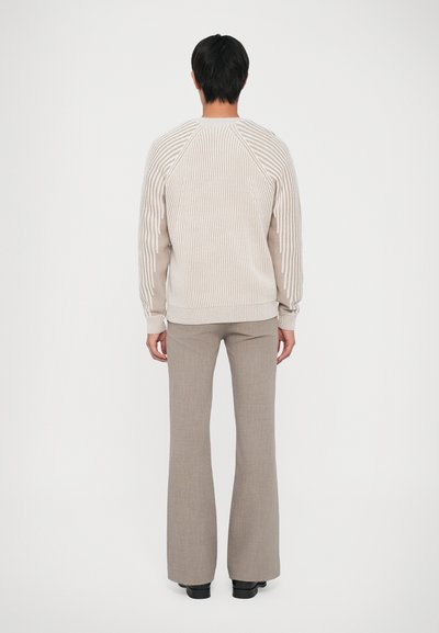 Man die met zijn rug naar de kijker staat, gekleed in een beige geribde trui met een gestreept patroon en lichtbruine uitlopende broek, staande op een effen witte achtergrond.