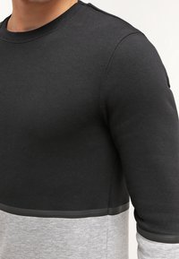 Hombre que lleva una sudadera de manga larga con bloques de color, con la parte superior negra y la parte inferior gris separadas por una delgada línea negra.