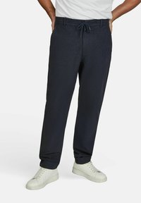 Marineblauwe broek van een gestructureerde stof, met een elastieken tailleband en trekkoord, en een taps toelopende pijp, gedragen met witte sneakers.