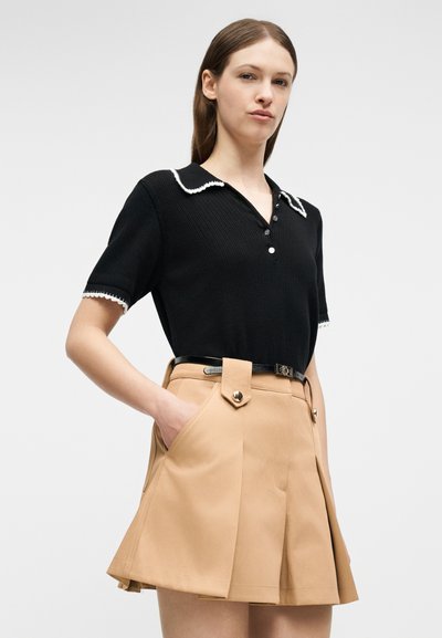 Νεαρή γυναίκα που φοράει μαύρο μπλουζάκι με κοντά μανίκια και λευκά περιθώρια, ενώ έχει μπεζ πλισέ σορτς ψηλόμεσο με μαύρη ζώνη, τα χέρια στις τσέπες.