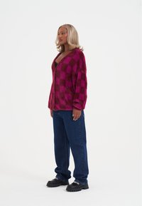 Chlupatý růžový kostkovaný kardigan s výstřihem do V, ve spojení s tmavě modrými džínami a robustními černými botami. Texturovaný povrch a volný střih.