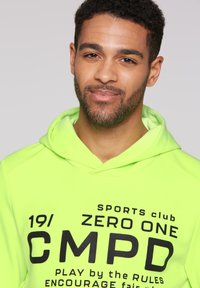 Felpa verde neon con testo stampato nero: "SPORTS club ZERO ONE CMPD GIOCA SECONDO LE REGOLE." Tessuto morbido con vestibilità rilassata e cappuccio con cordoncino.