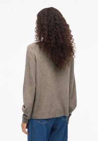 Ženska z dolgimi kodrastimi lasmi, oblečena v sivo pleteno pulover in modre kavbojke, stoji obrnjena proti belini.