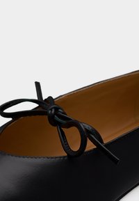 Black leather ballet flat with a small decorative bow on top and tan interior lining.