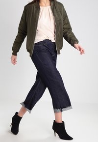 Giacca bomber verde oliva su una maglietta rosa chiaro, abbinata a jeans scuri a gamba larga con orli sfrangiati e stivaletti neri.
