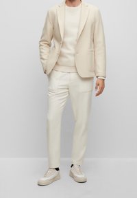 Beige Wollblazer mit zwei Fronttaschen, kombiniert mit einem passenden beige gestrickten Pullover und maßgeschneiderten Hosen. Abgerundet mit hellen Sneakers.