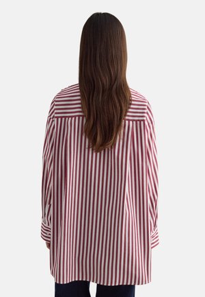 Person mit langen braunen Haaren, die ein locker sitzendes, rot-weiß gestreiftes Hemd mit vertikalen Streifen trägt und mit dem Rücken zum Betrachter vor einem schlichten Hintergrund steht.