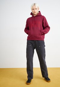 Κρασί φούτερ με μεγάλο λογότυπο "OBEY" σε κόκκινο χρώμα, με μπροστινή τσέπη και κουκούλα με κορδόνι, συνδυασμένο με σκούρα παντελόνια και μαύρα παπούτσια.