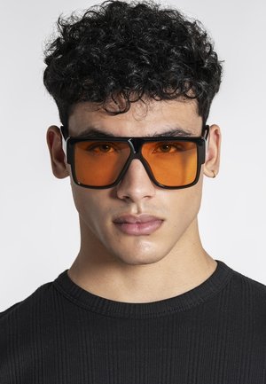 Mladić s kovrčavom crnom kosom nosi velike kvadratne crne sunčane naočale s narančastim staklima i crnu rebrastu majicu.