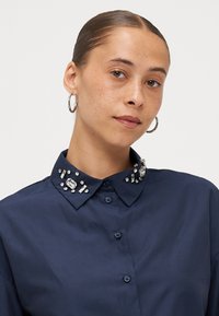 Navy blue shirt featuring a jeweled collar with clear and textured embellishments, button front, and a smooth fabric texture.